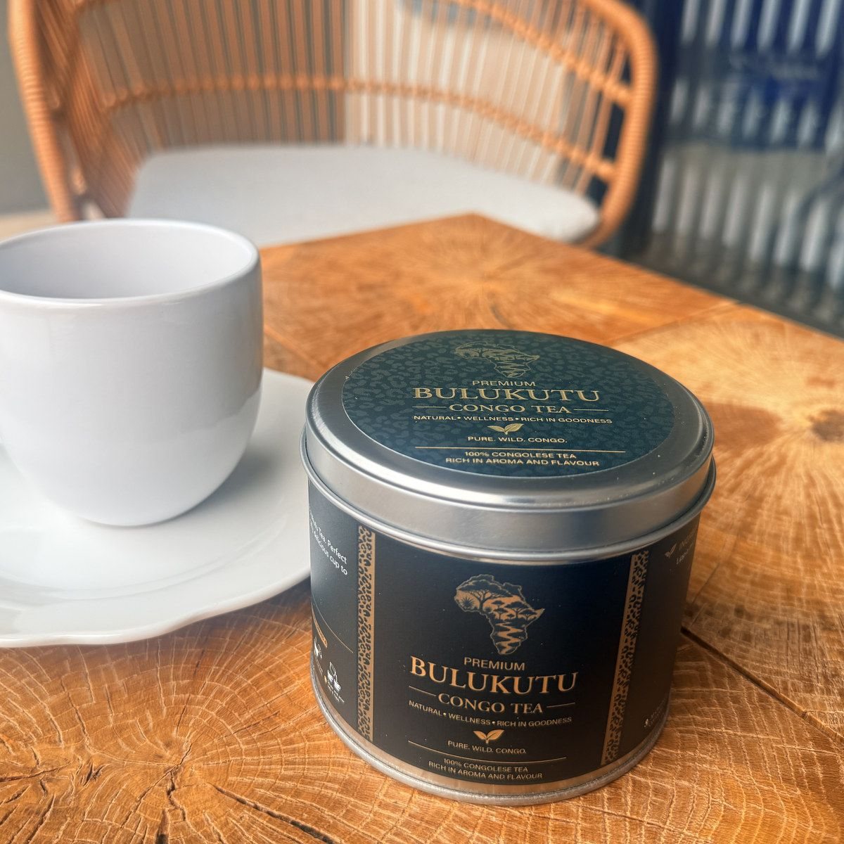 The Bulukutu tin beside a white cup and saucer on a wooden table