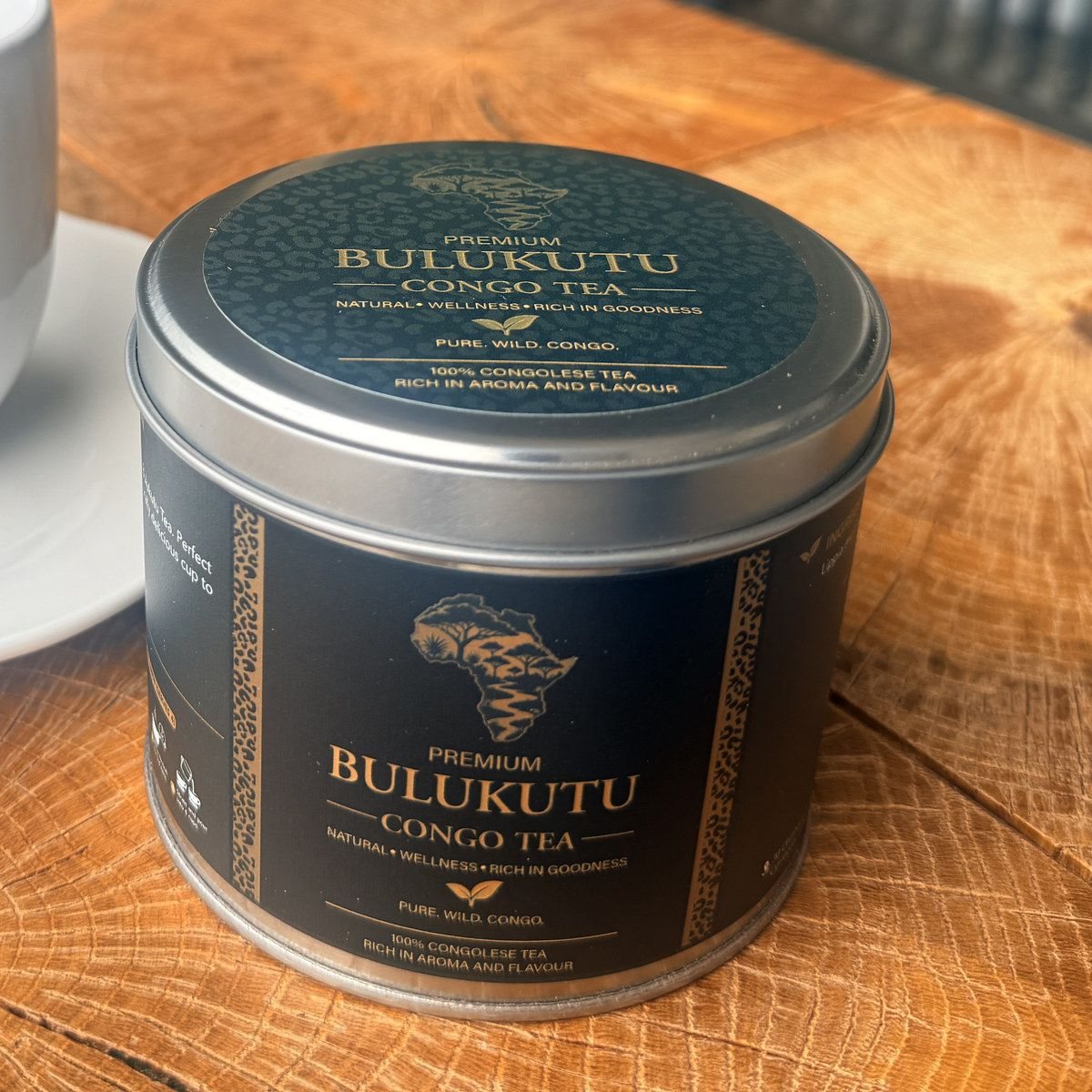 A tin of Premium Bulukutu Congo Tea on a wooden table, its teal lid printed in gold with the outline of Africa